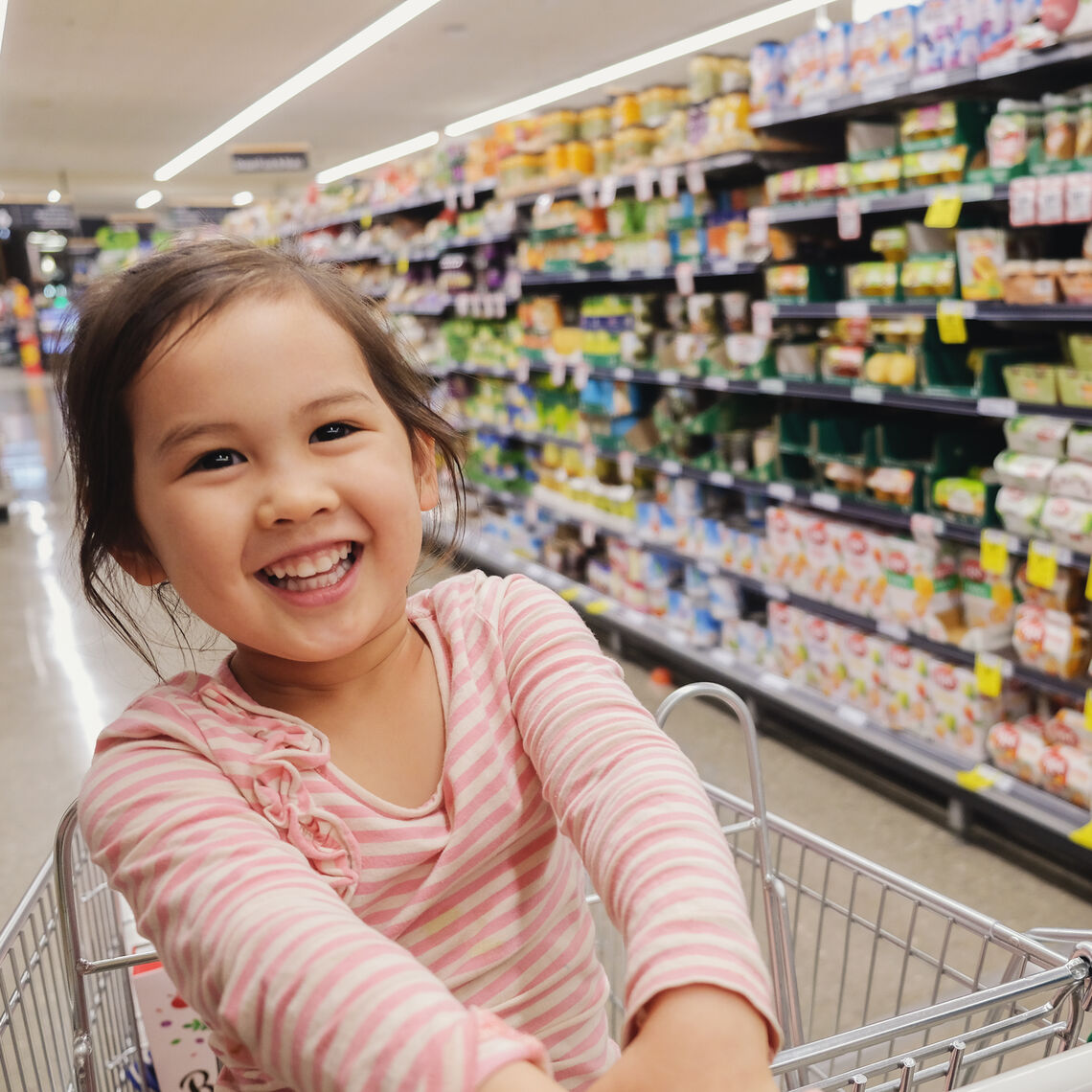 Happy little multiethnic girl sitting in a trolley, shopping cart at supermarket, grocery store
