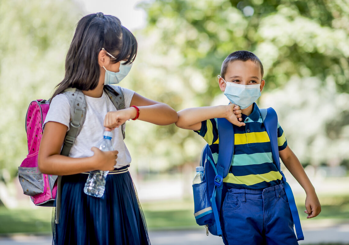 A young boy and girl wearing masks, bumping elbows outside with backpacks on