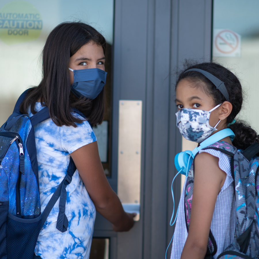 Two middle school aged girls in masks are wearing backpacks, looking over their shoulders at camera walking into front doors