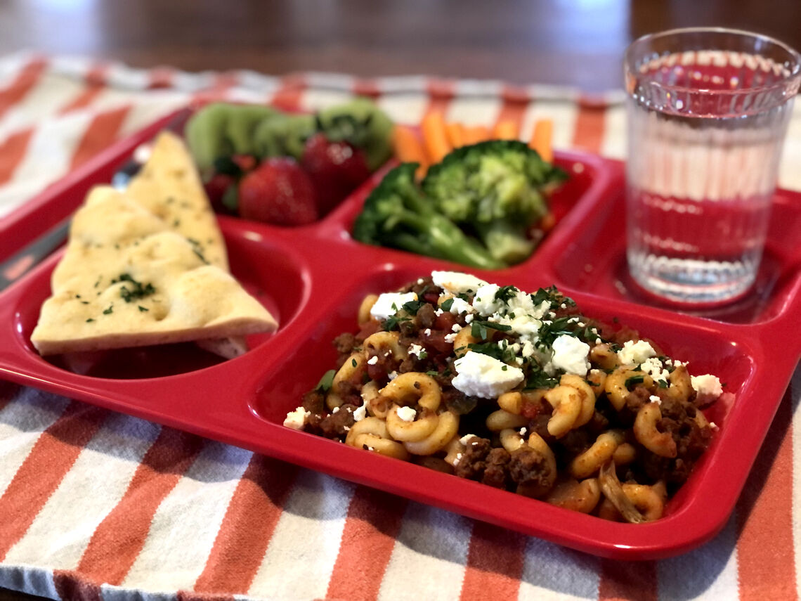 A red school lunch tray with Project Bread school lunch recipe, Amy Greek's Chop Suey served with pita, broccoli, and a fresh side of fruit