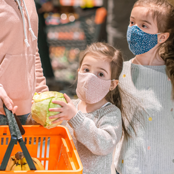 Two sisters with face masks help mom grocery shopping, adding a fresh head of lettuce to the basket