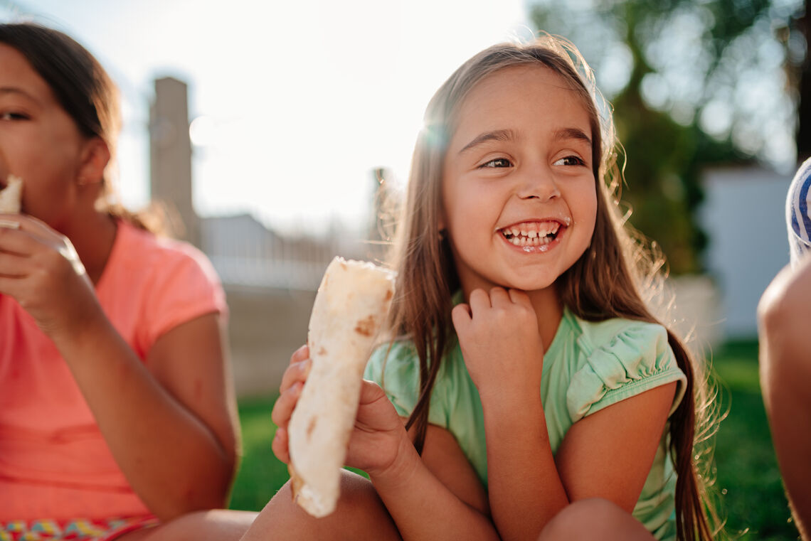 Girls sitting outdoor on the ground at backyard eating sandwiches and smiling