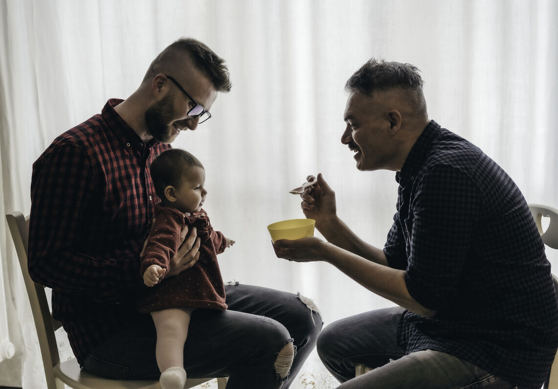 Two dads feed their baby girl in the kitchen