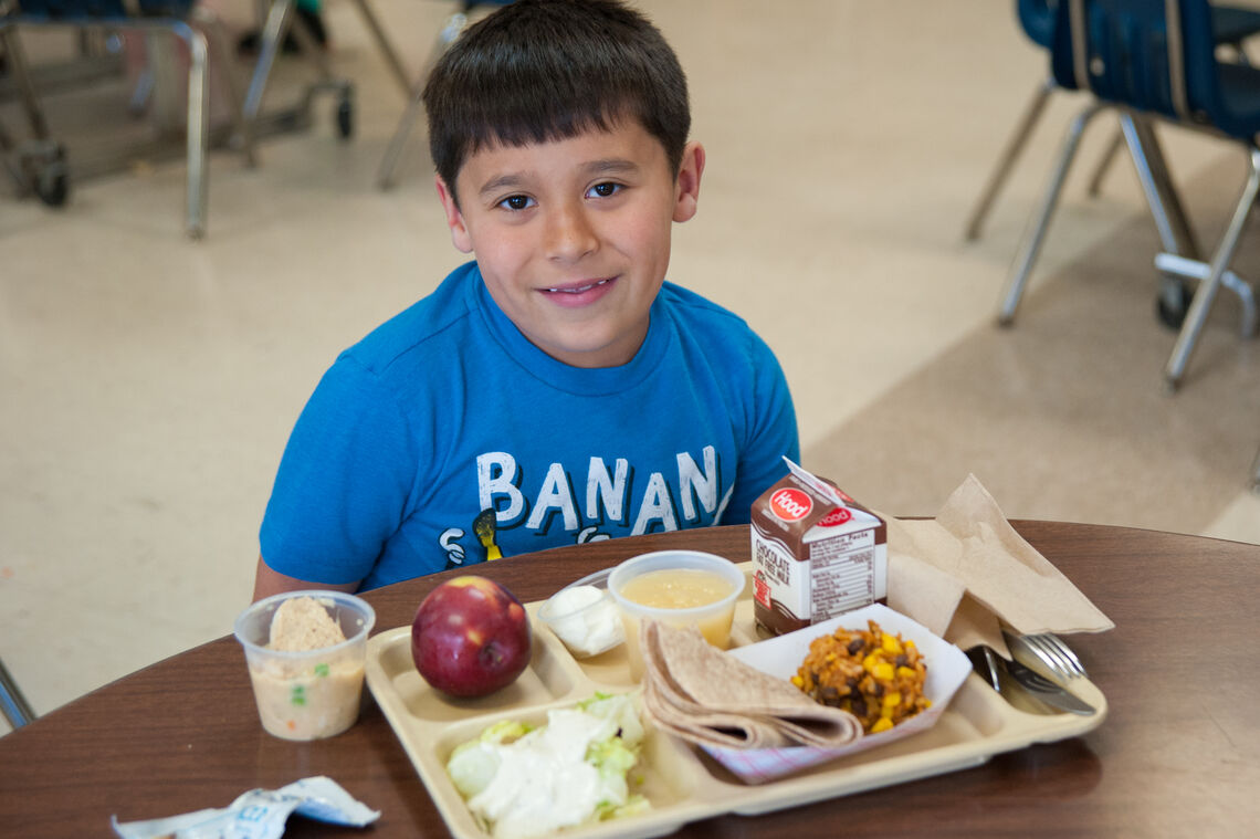 Young Latino boy smiling at school cafeteria table with a tray of black bean burrito lunch