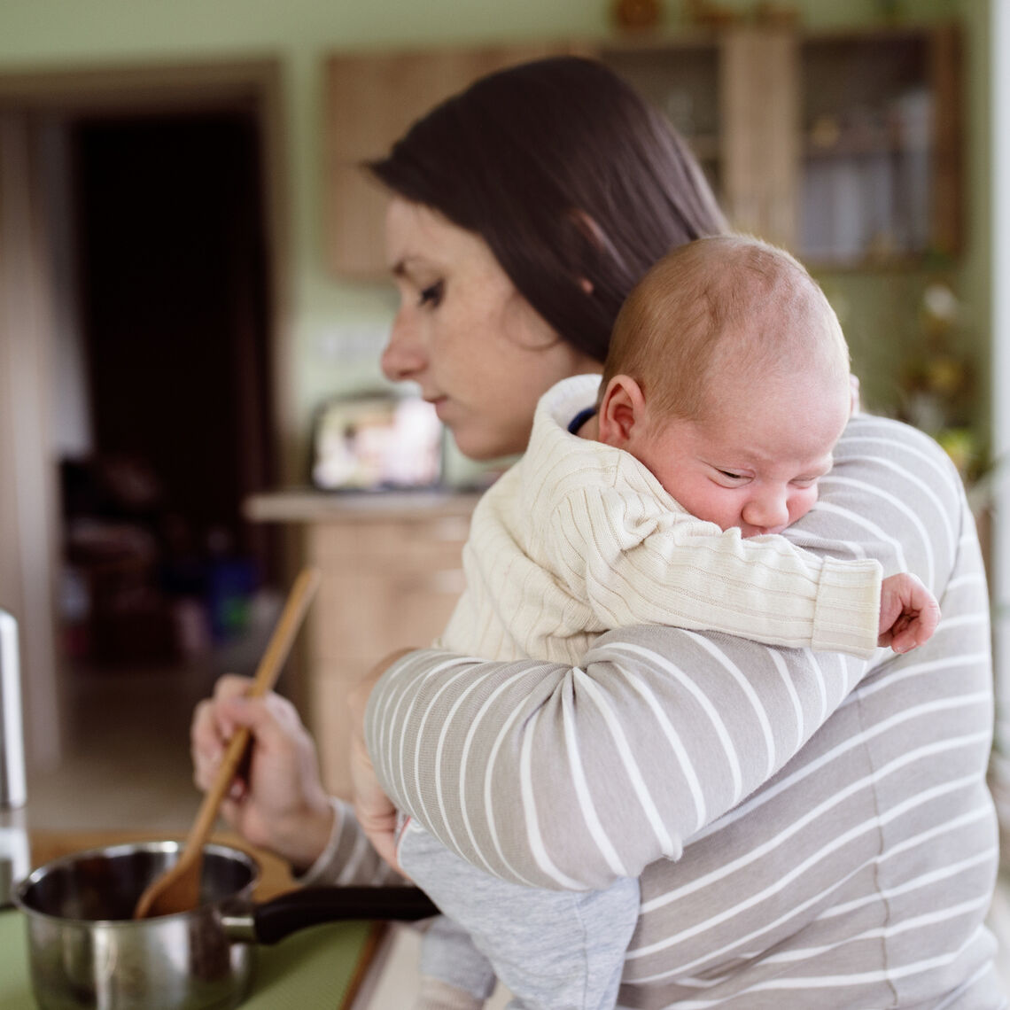 Beautiful young mother at home in the kitchen holding her newborn baby son, cooking, mixing something in pan (Beautiful young mother at home in the kitchen holding her newborn baby son, cooking, mixing something in pan.