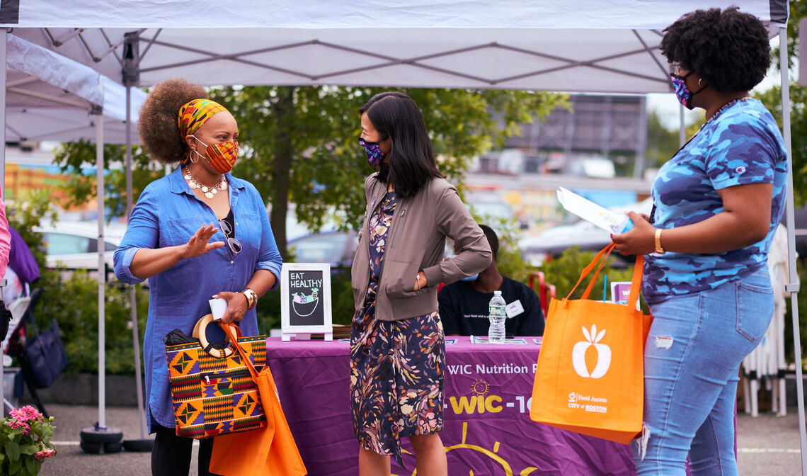 Three women in vibrant clothing standing in front of a WIC table at the Mattapan Farmer's Market