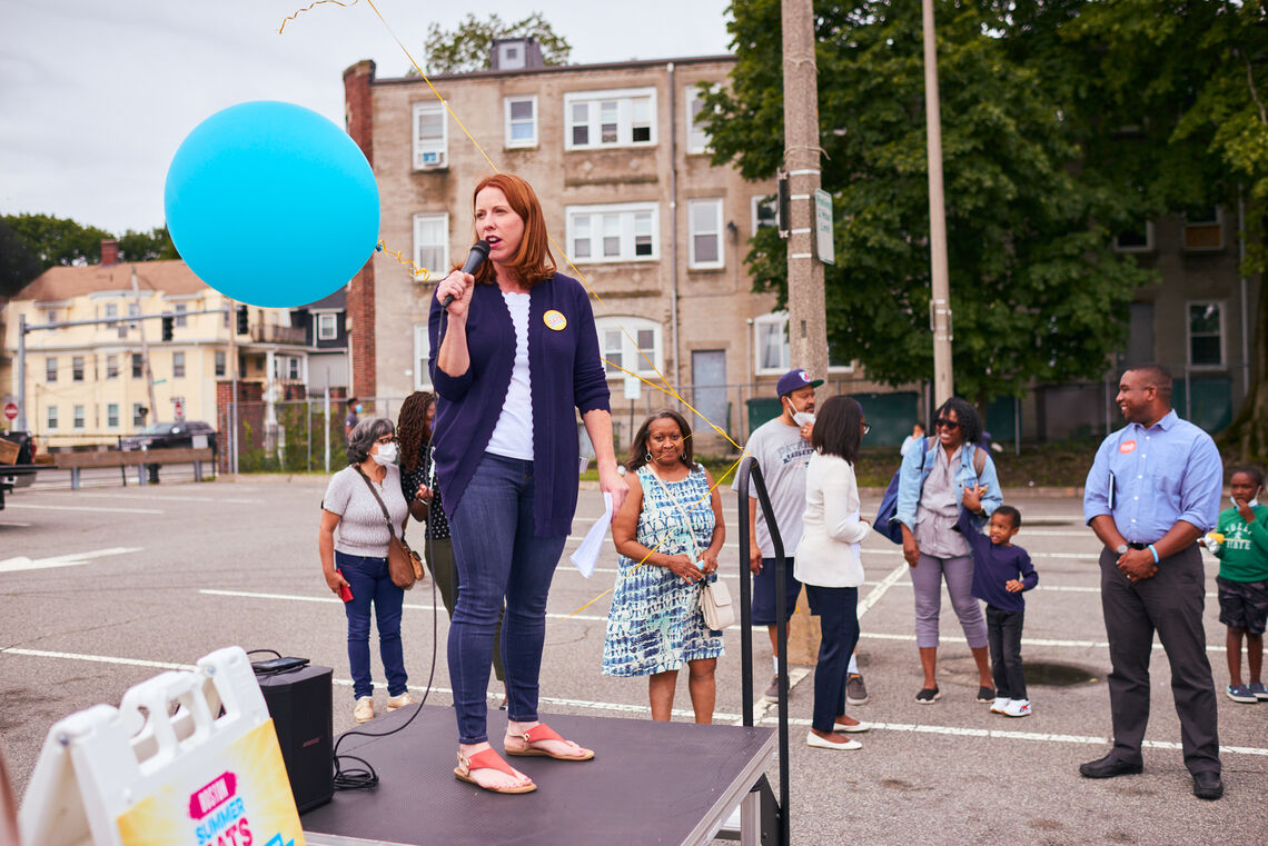 Erin Mcaleer stands on a platform, speaks to a crowd at opening day of Mattapan Food & Fitness Coalition farmers market
