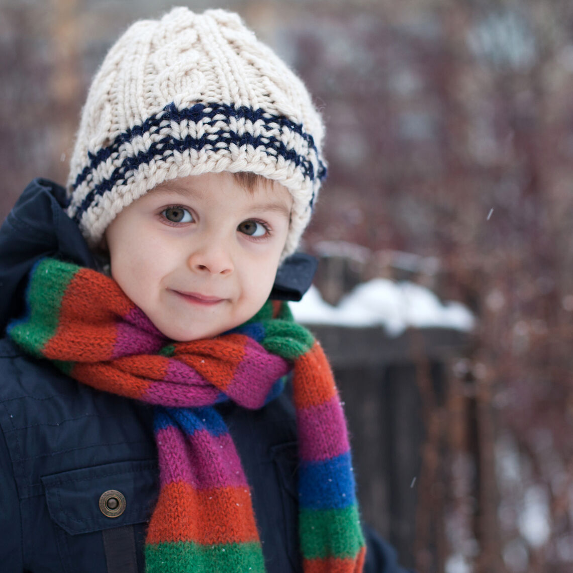 Young boy dressed in winter hat, scarf, and coat, outdoors smiling at the camera
