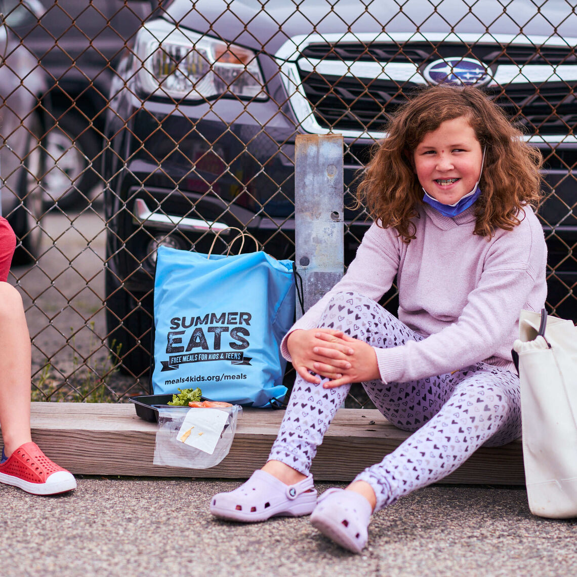 A young boy and girl lean up against a fence at a farmers market enjoy their Summer Eats meal