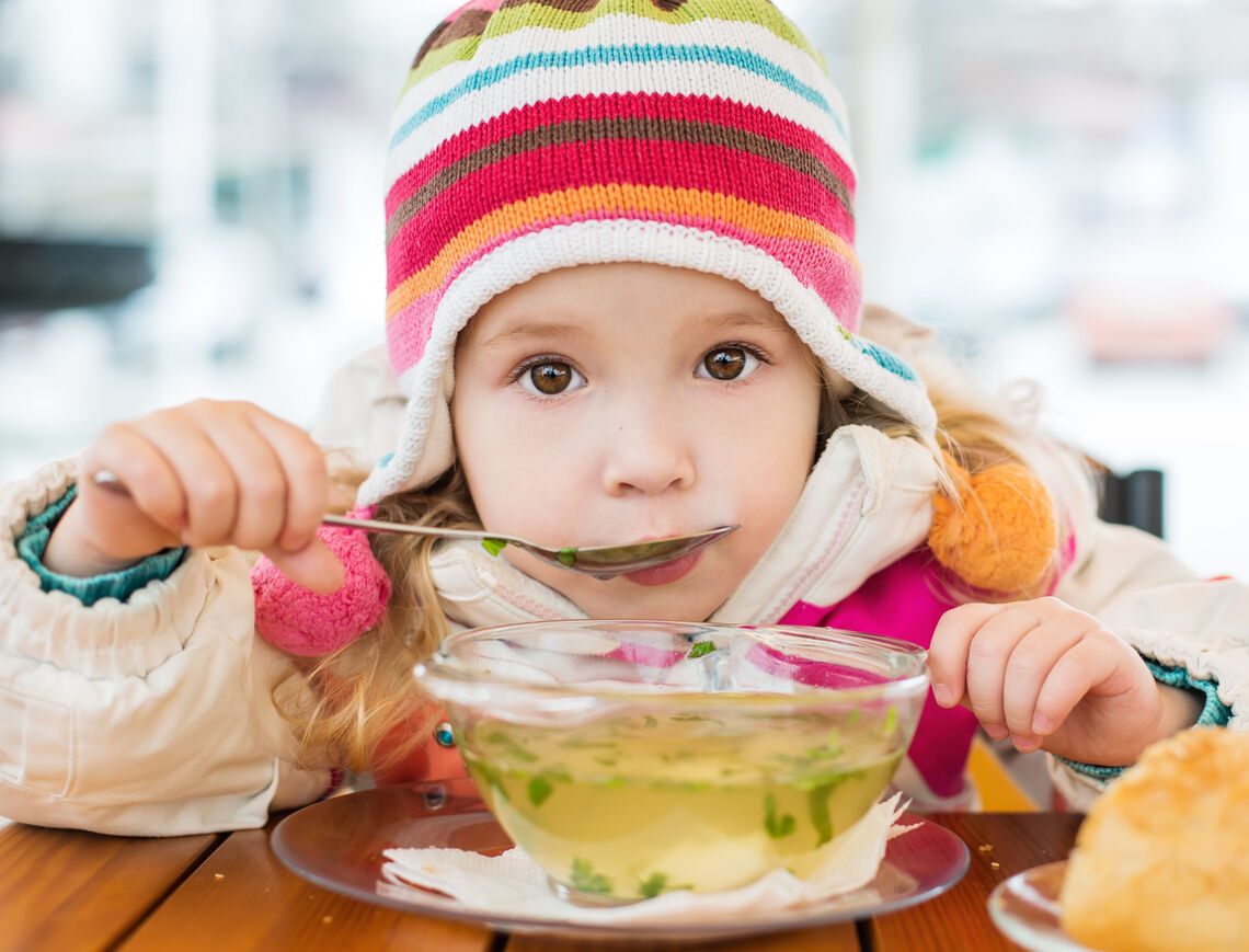 Young girl wearing winter clothes sits at a table eating hot soup with a spoon to her mouth