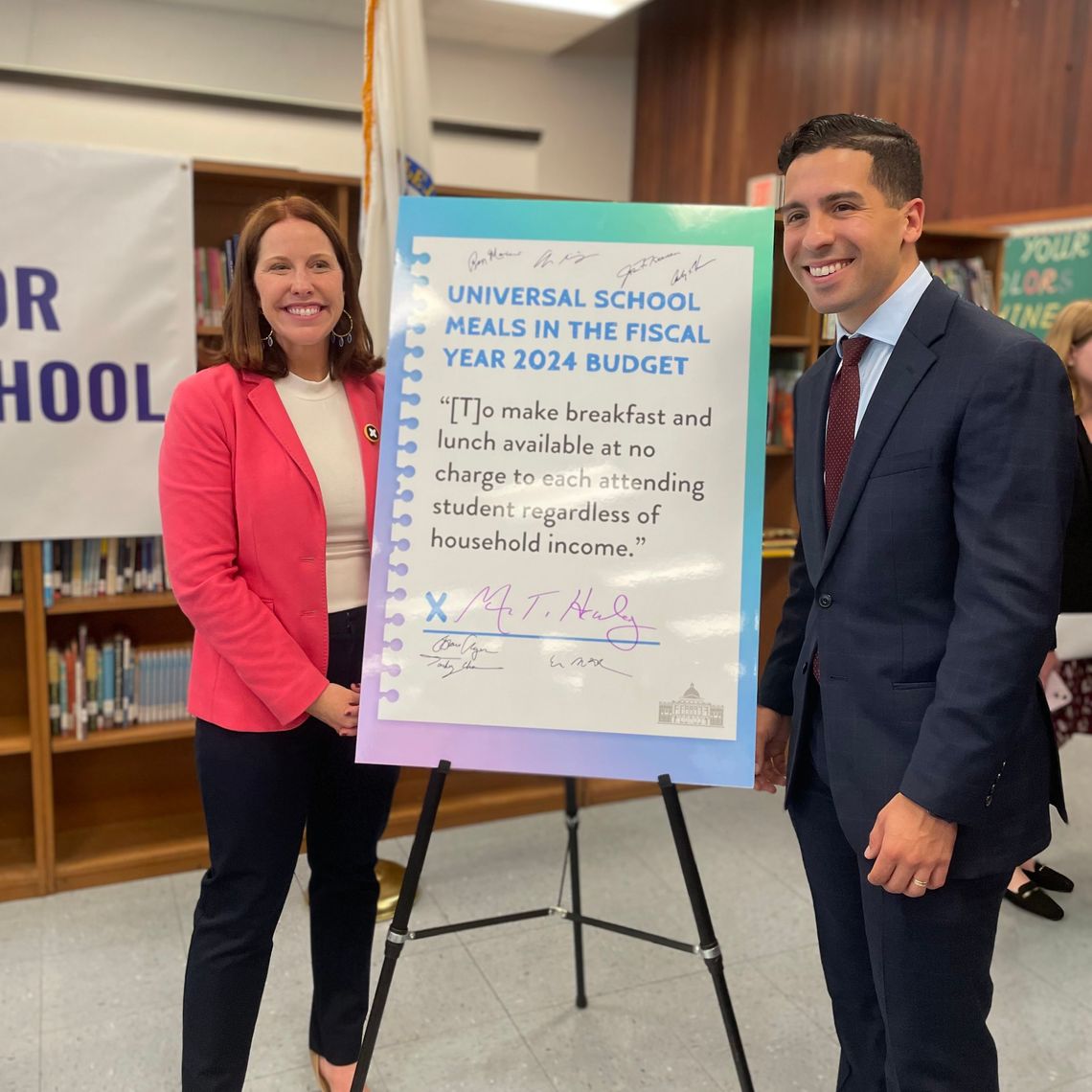 Erin McAleer, President and CEO of Project Bread stands along with Representative Andres X. Vargas, next to a large poster advertising Universal Free School Meals featuring Governor Maura Healey's signature