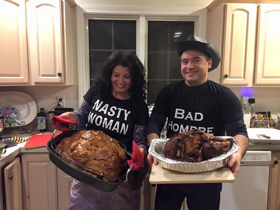 Adriana Mendes-Sheldon stands smiling next to her husband, both are holding roasted turkeys on large trays.